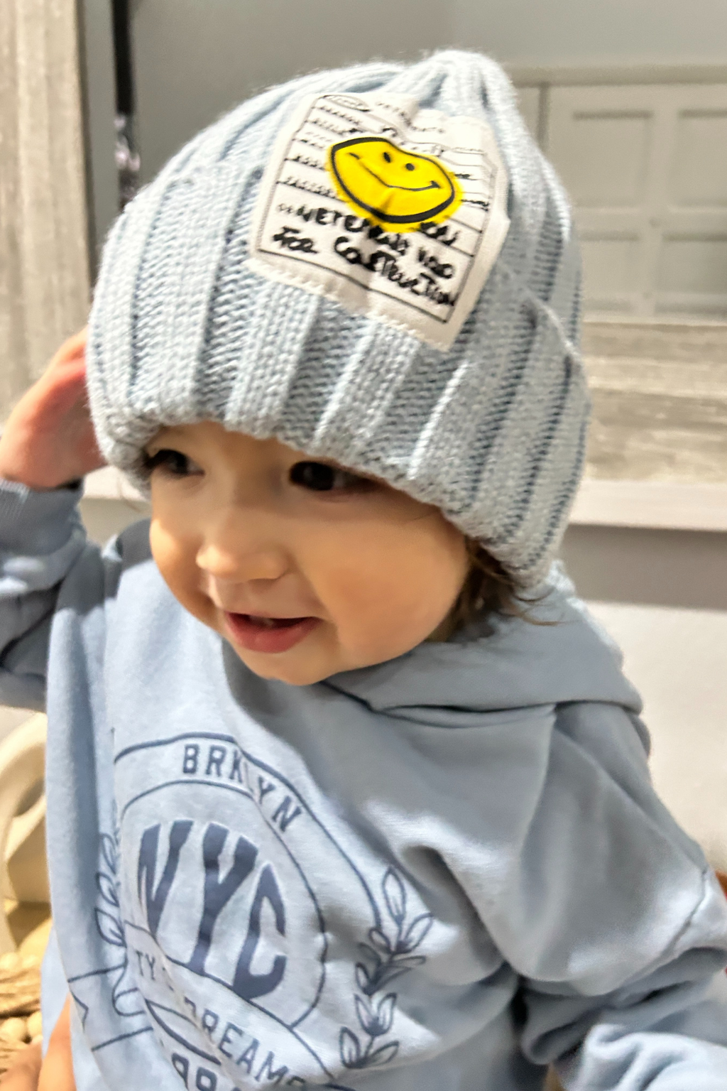 A close-up of a toddler wearing a light blue ribbed knit beanie with a white patch featuring a yellow smiley face and text, along with a matching light blue hoodie.