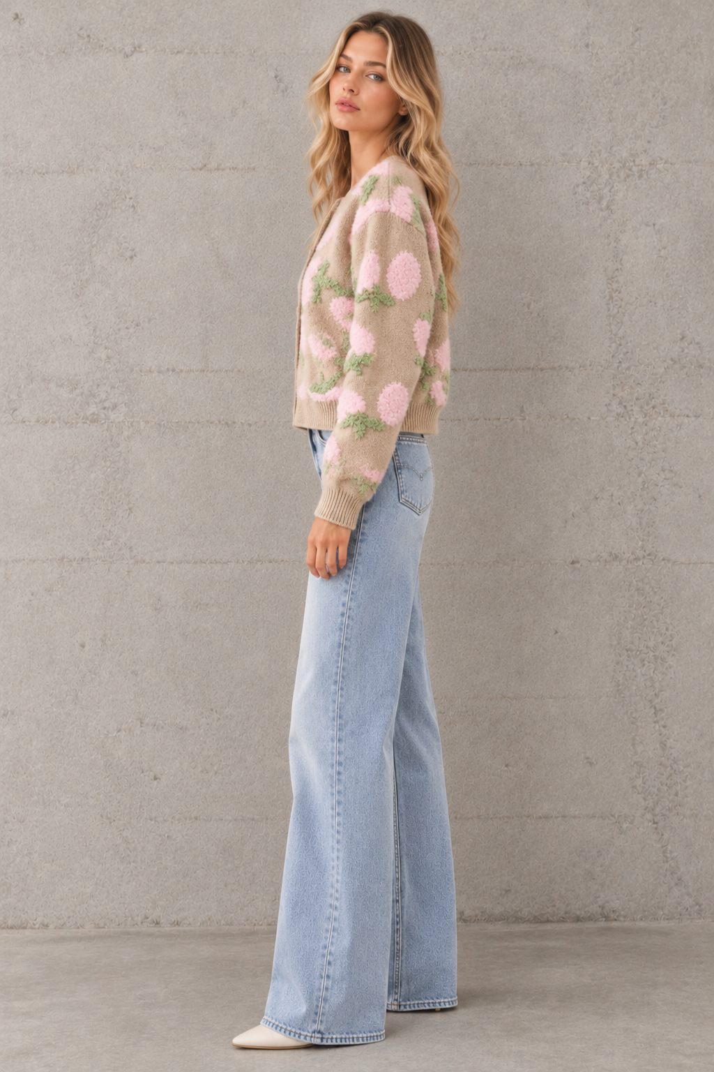 A woman stands in profile wearing a light beige knit cardigan with large pink and green floral motifs, paired with light-wash wide-leg jeans and white pointed shoes, against a textured gray concrete wall.