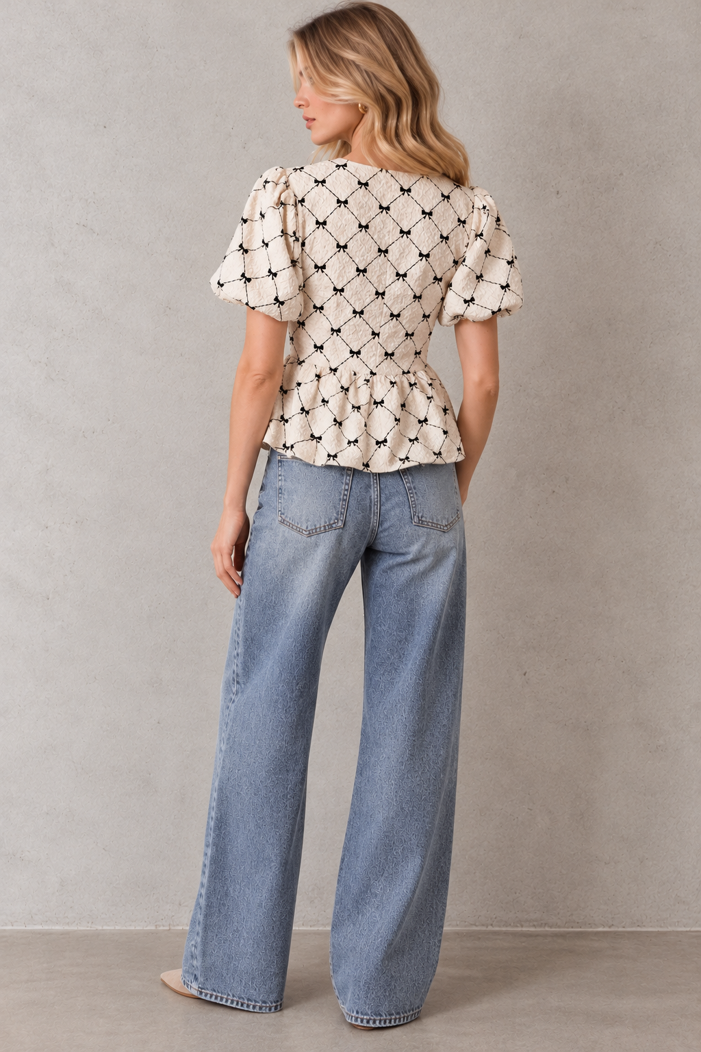 A woman is shown from behind wearing a cream-colored short-sleeve peplum top with a black trellis pattern and light-wash blue wide-leg jeans, standing against a textured gray wall.