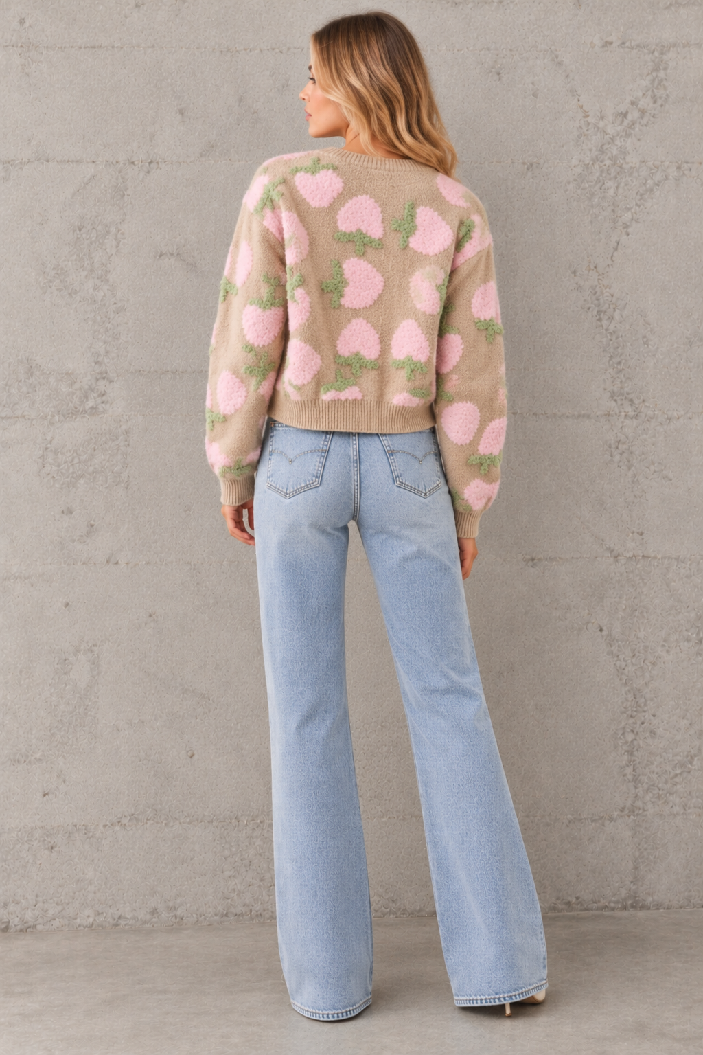 A woman viewed from the back wearing a beige knit sweater with a pink and green strawberry pattern, paired with light blue wide-leg jeans, standing against a textured gray concrete wall.