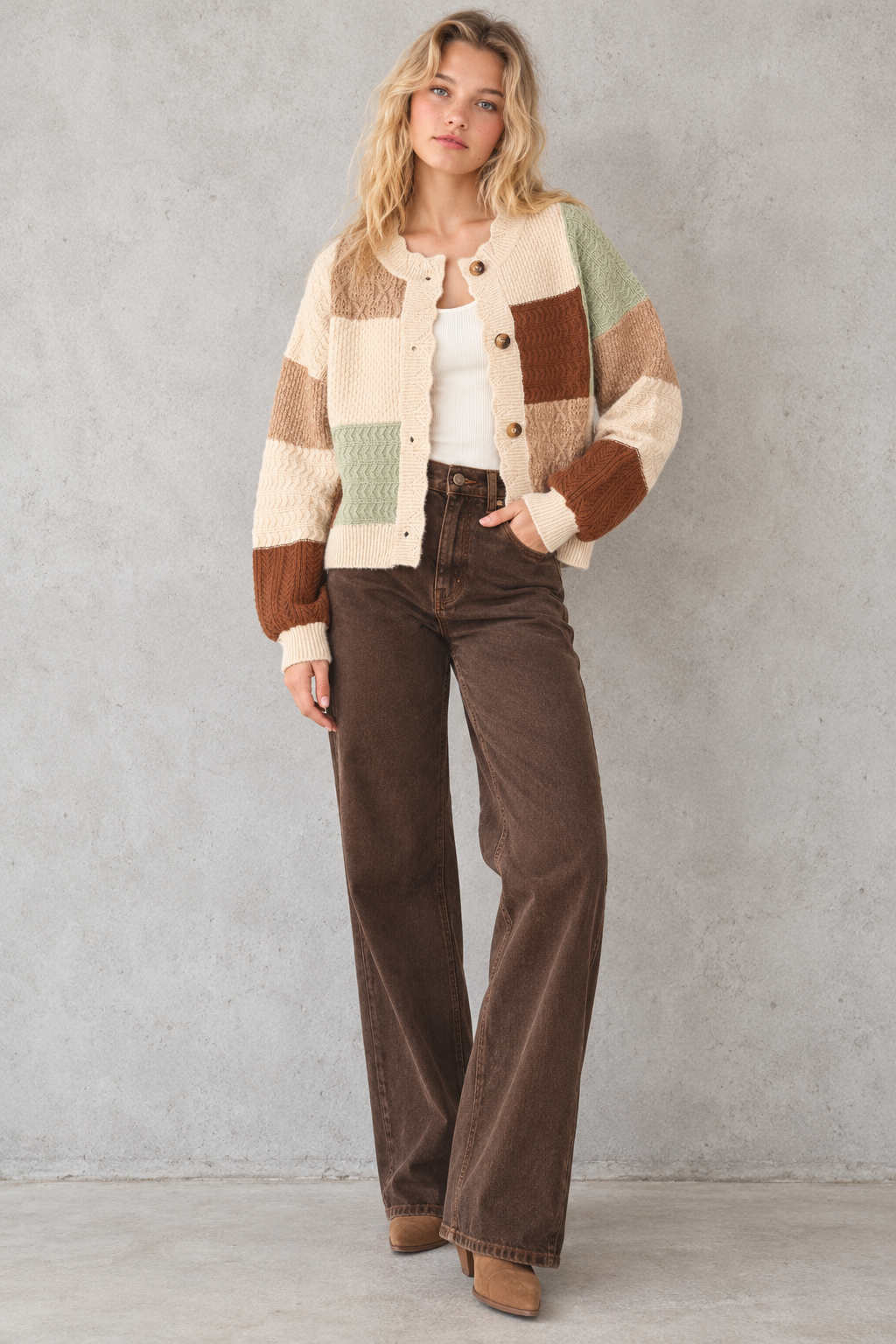A woman stands against a textured light gray wall wearing a cream, brown, and sage green patchwork knit cardigan, brown wide-leg jeans, and tan boots.