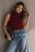 A woman wearing a sleeveless ribbed turtleneck sweater in deep burgundy, paired with high-waisted wide-leg blue jeans and a dark brown shoulder bag, posing against a textured concrete wall.