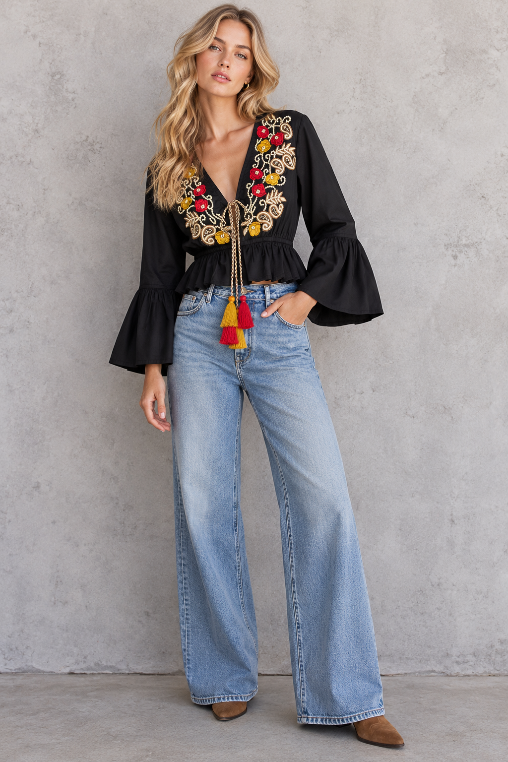 A woman modeling a black long-sleeve blouse with colorful floral embroidery and tassel ties, paired with light blue wide-leg jeans and brown shoes, standing against a textured gray wall.