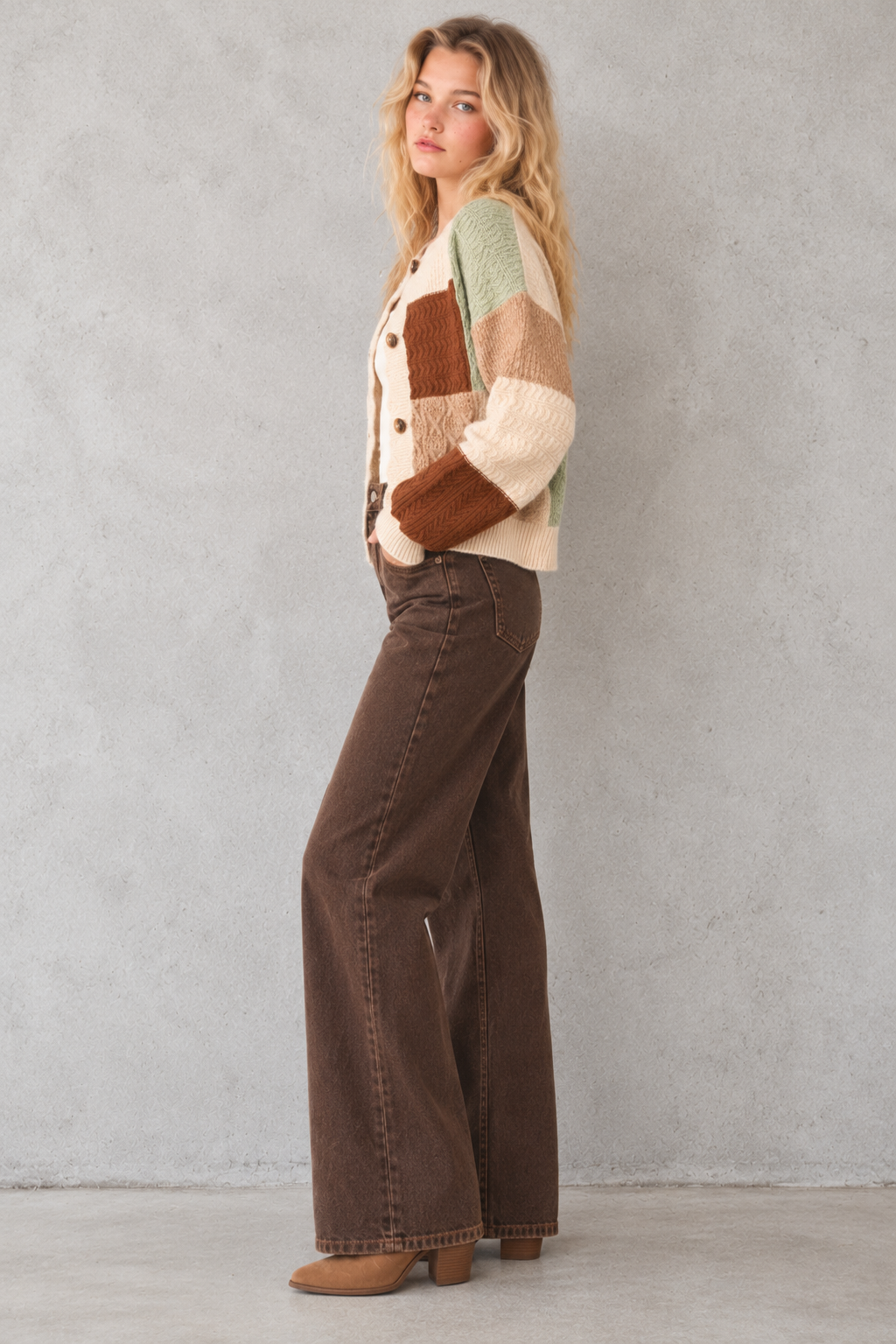 A woman stands in profile wearing a multicolor patchwork knit cardigan with brown suede-like panels, dark brown wide-leg jeans, and tan heeled boots against a textured gray wall.
