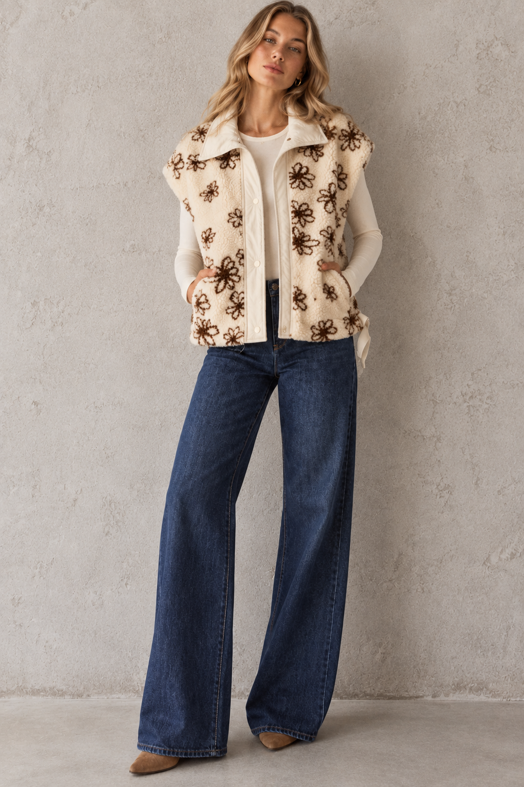 A woman models a sleeveless cream-colored vest with brown floral embroidery, worn over a white top and paired with wide-leg blue jeans, standing against a textured light gray wall.
