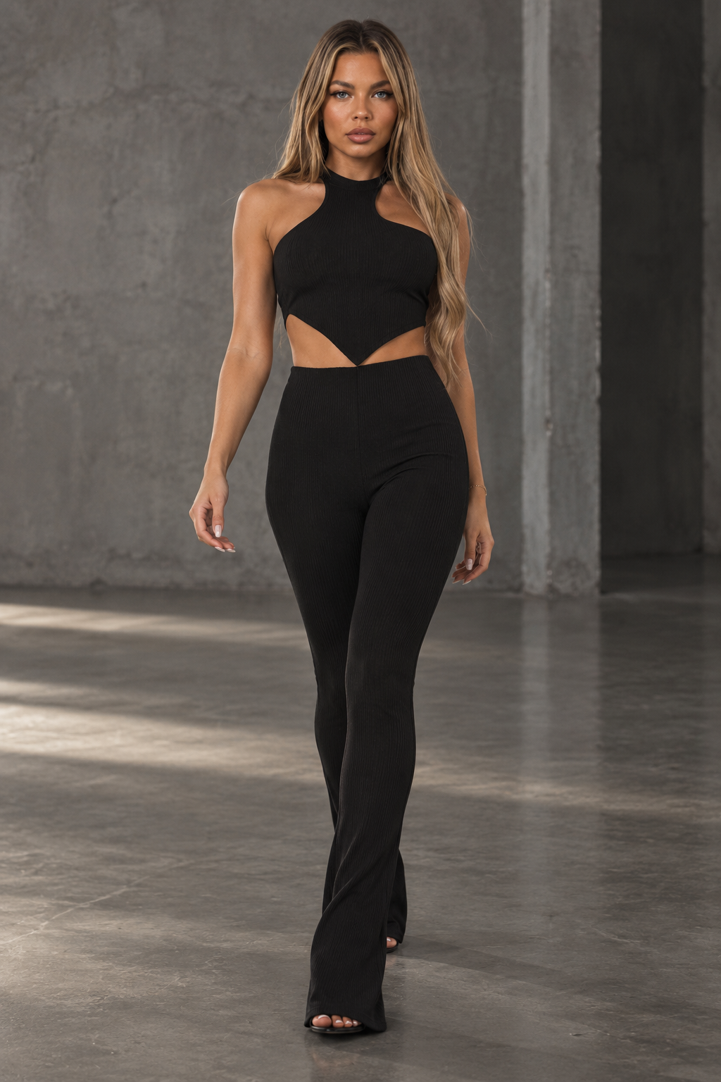 A woman modeling a black two-piece cutout outfit consisting of a halter-style crop top and matching high-waisted flared leggings, photographed in a concrete industrial interior.