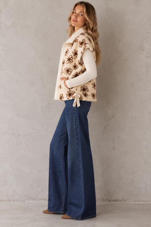 A woman stands in profile wearing a cream sleeveless vest with brown floral embroidery, a white long-sleeve top, and wide-leg blue jeans against a textured light gray wall.