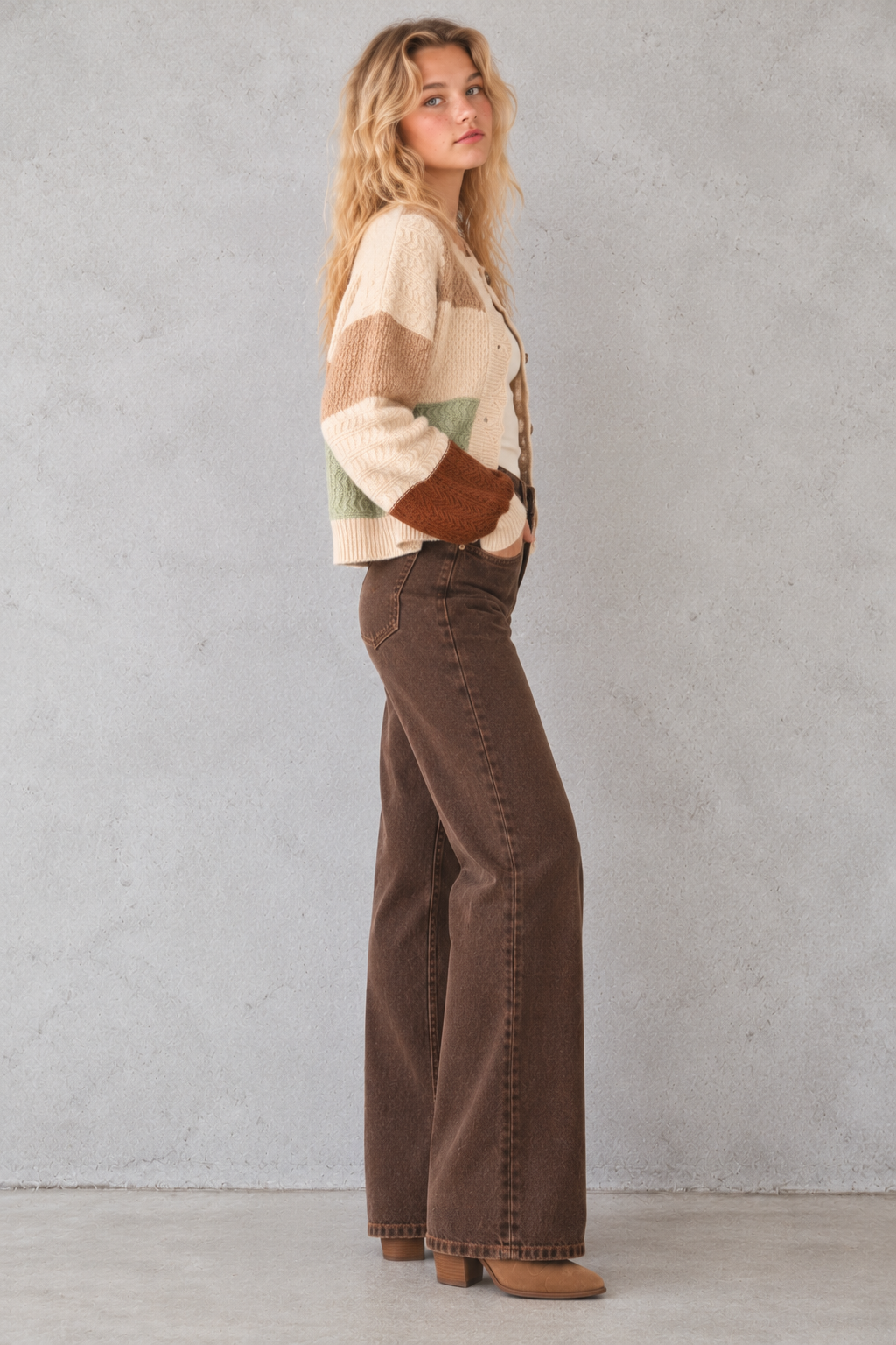 A woman stands in profile wearing a cream, tan, and brown color-block knit cardigan with brown suede-like cuffs, paired with dark brown wide-leg jeans and tan boots, against a textured light gray wall.