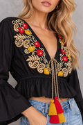A close-up of a woman wearing a black long-sleeve crop top with a deep V-neck, decorated with colorful floral and paisley embroidery in red, yellow, and gold, featuring a drawstring tie with red and yellow tassels, paired with blue jeans.