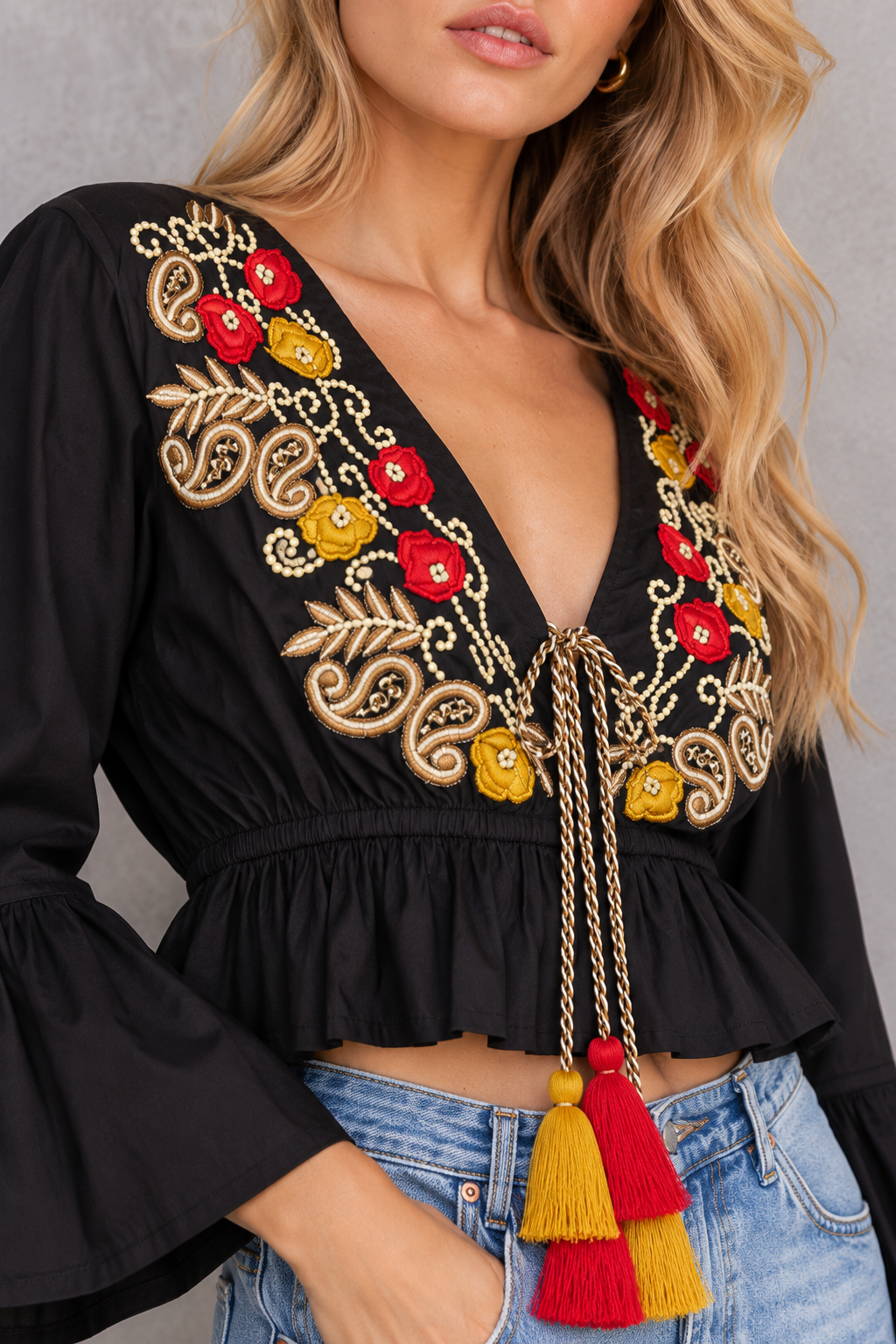 A close-up of a woman wearing a black long-sleeve crop top with a deep V-neck, decorated with colorful floral and paisley embroidery in red, yellow, and gold, featuring a drawstring tie with red and yellow tassels, paired with blue jeans.