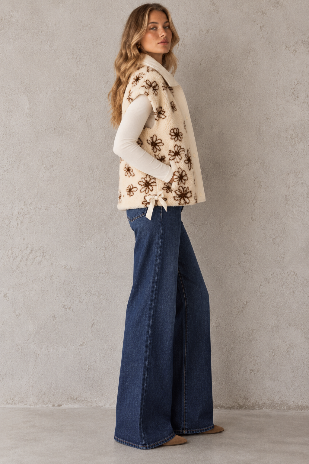 A woman stands in profile wearing a cream sleeveless vest with a brown floral pattern over a white long-sleeve top and dark blue wide-leg jeans, against a textured light gray wall.