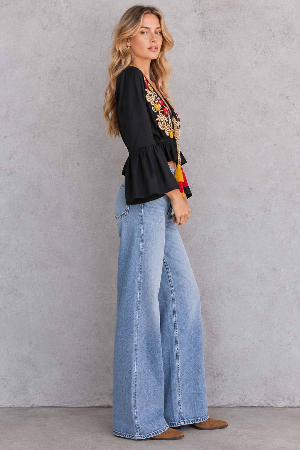 A woman stands in profile against a textured gray wall, wearing a black long-sleeve blouse with colorful floral embroidery on the chest and wide-leg light blue jeans.