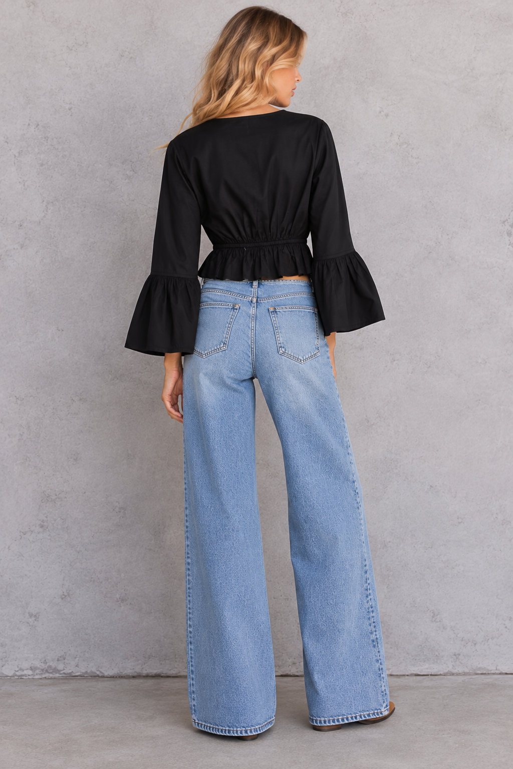 A woman shown from the back wearing a black long-sleeve crop top with flared bell sleeves and light-wash wide-leg denim jeans, standing against a textured gray wall.