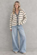A woman stands against a textured gray wall wearing a cream and gray striped fuzzy knit cardigan with wide-leg light blue jeans and beige shoes.