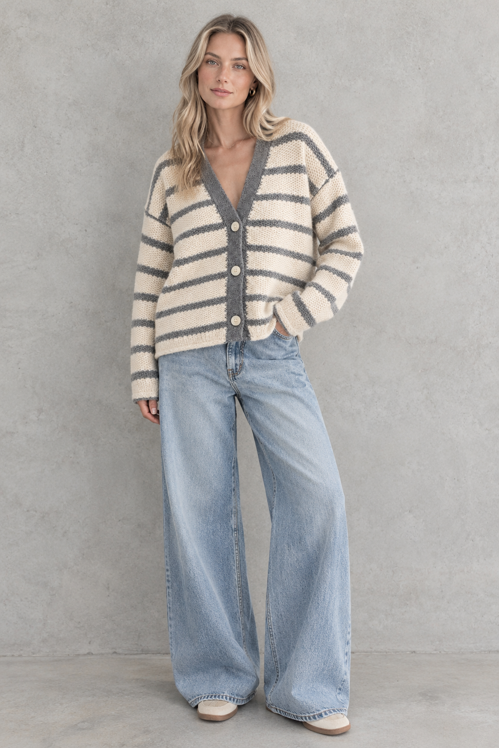 A woman stands against a textured gray wall wearing a cream and gray striped fuzzy knit cardigan with wide-leg light blue jeans and beige shoes.