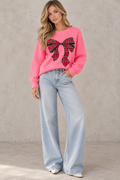 A woman modeling a bright pink crewneck sweater with a large red, black, and green plaid bow graphic on the front, paired with light-wash wide-leg jeans against a textured gray wall.