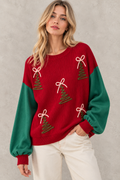 A woman wearing a festive Christmas sweater with a red body, green sleeves, and embroidered green Christmas trees with white bows, paired with off-white pants, set against a textured gray wall.