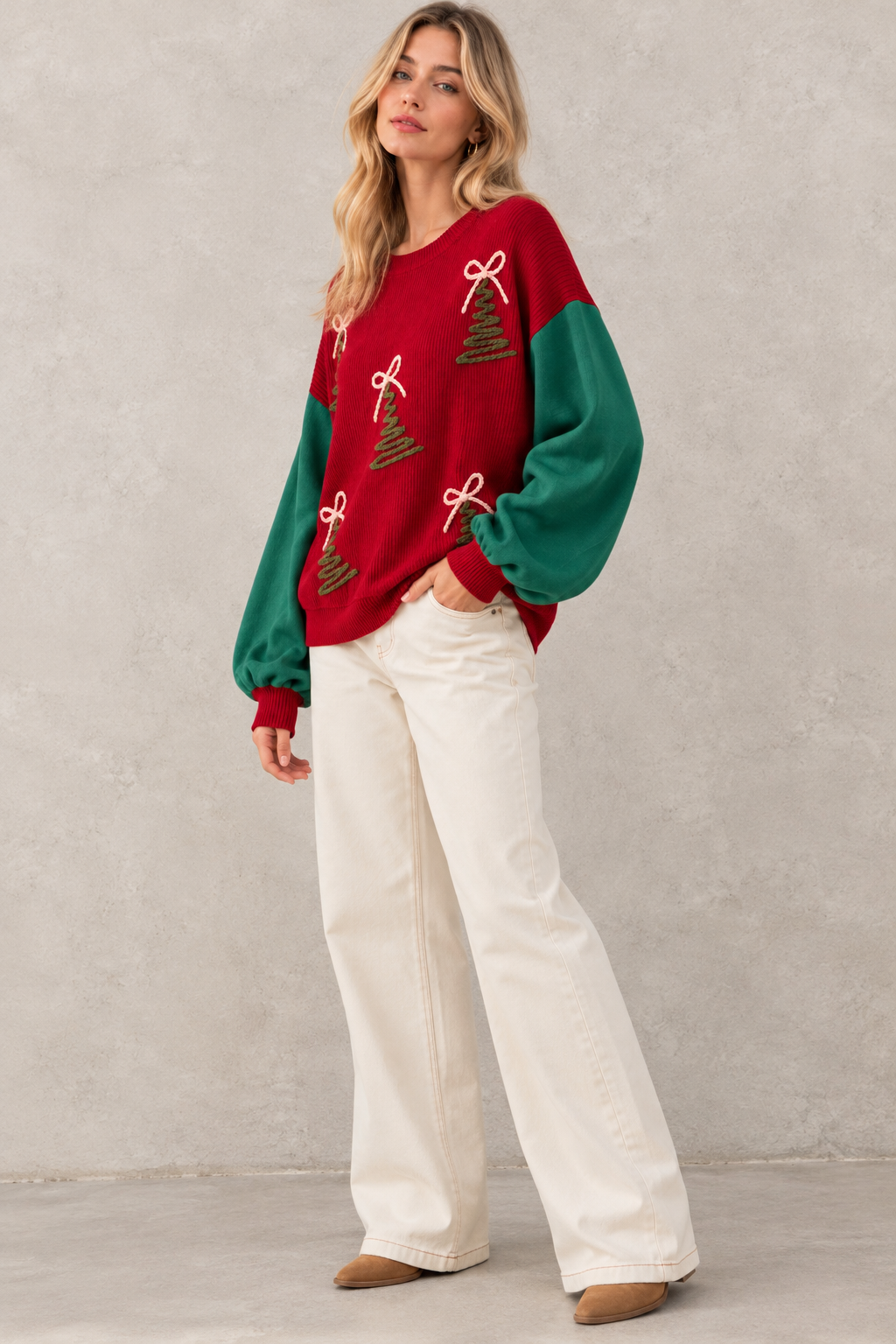 A woman models a festive Christmas-themed crewneck sweater with red and green color-blocked sleeves and embroidered pine trees and bows, paired with off-white wide-leg jeans and tan shoes, set against a textured gray wall.