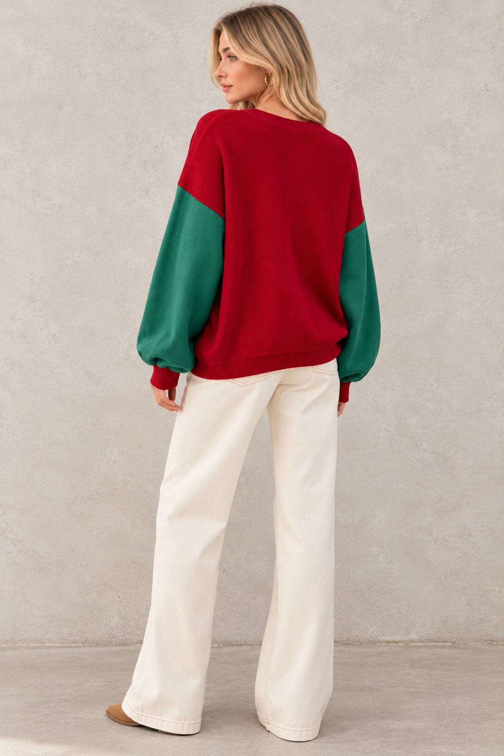 A woman wearing a color-block knit sweater with a red body and green sleeves, paired with cream wide-leg pants, standing against a textured light gray wall.