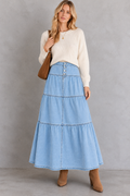 A woman stands against a textured gray studio wall wearing a cream ribbed knit sweater, a light blue tiered denim skirt with button details, and brown ankle boots, while carrying a brown shoulder bag.