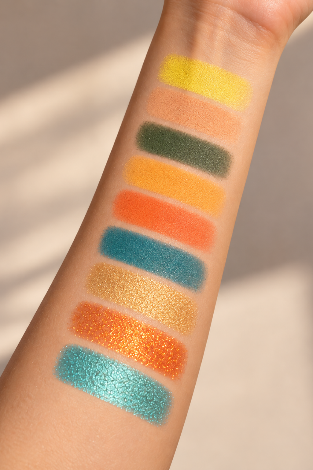 A close-up of a forearm with eight horizontal eyeshadow swatches in various colors and finishes, including matte yellow, peach, olive green, orange, bright orange, teal blue, shimmery gold, glitter orange, and sparkling teal, set against a softly lit beige background.