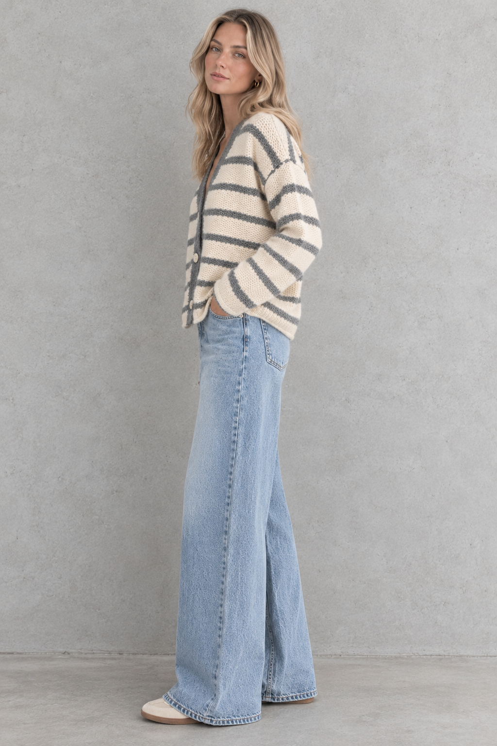 A woman stands in profile wearing a cream knit cardigan with dark gray horizontal stripes, light-wash wide-leg denim jeans, and beige shoes against a textured gray studio wall.