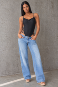 A woman modeling a black satin slip-style bodysuit with thin spaghetti straps, side cutouts, and light-wash wide-leg jeans, standing against a textured gray concrete wall.