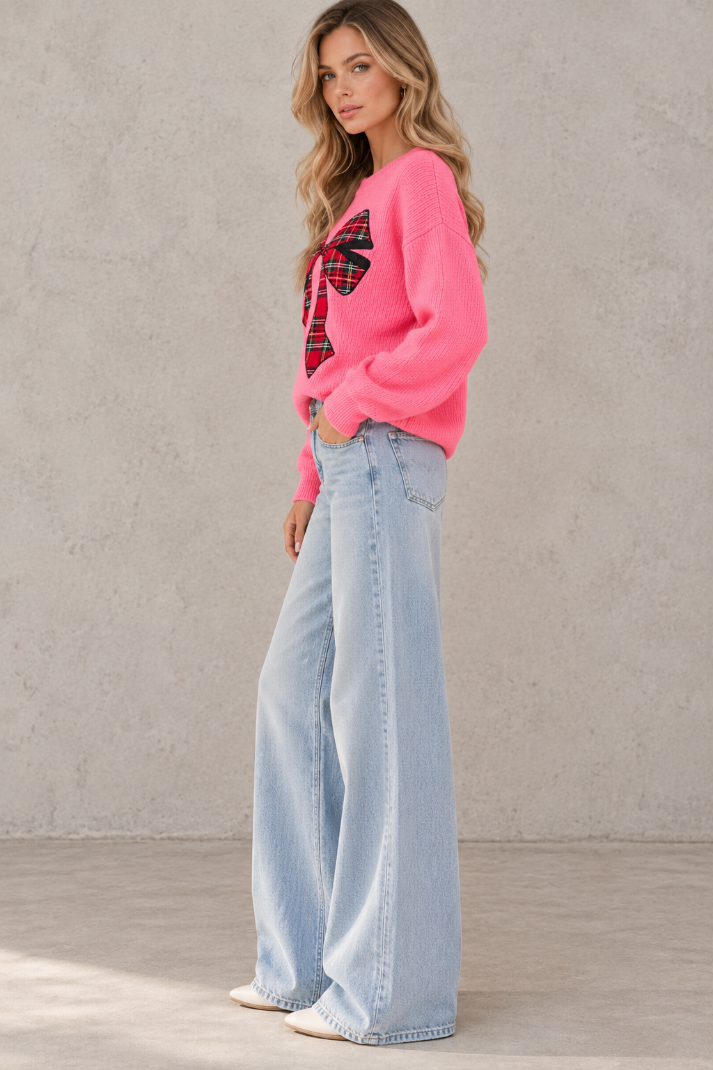 A woman stands in profile wearing a bright pink knit sweater with a red and black plaid bow graphic on the chest, paired with light-wash wide-leg jeans and white sneakers, against a textured light gray wall.