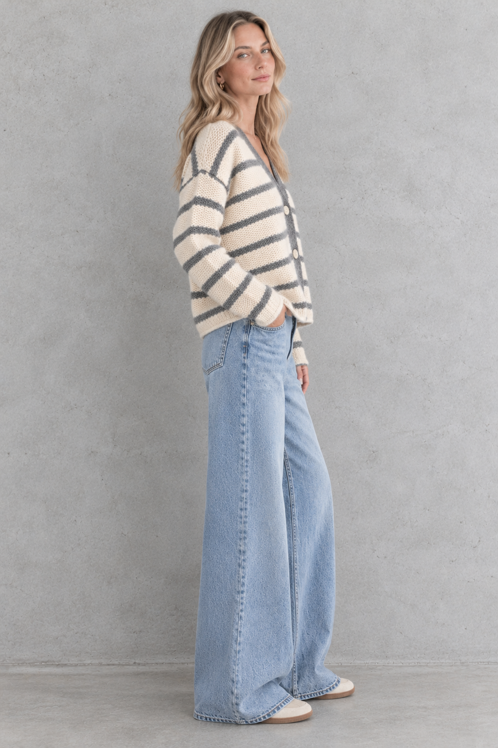 A woman stands in profile wearing a cream knit cardigan with dark gray horizontal stripes and light-wash wide-leg jeans against a textured gray wall.