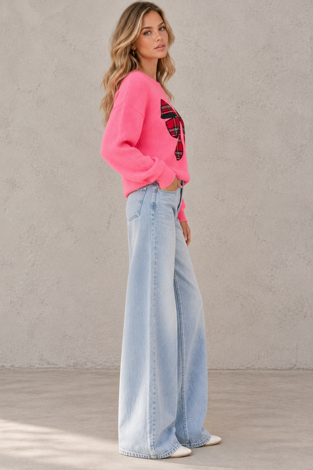 A woman modeling a bright pink cropped sweater with a red and black graphic print on the front, paired with light-wash wide-leg jeans and white sneakers, standing against a textured gray wall.