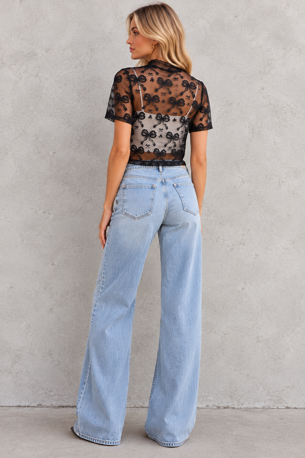 A woman stands with her back to the camera wearing a sheer black floral lace short-sleeve crop top over a white camisole, paired with light-wash wide-leg jeans, against a textured gray wall.