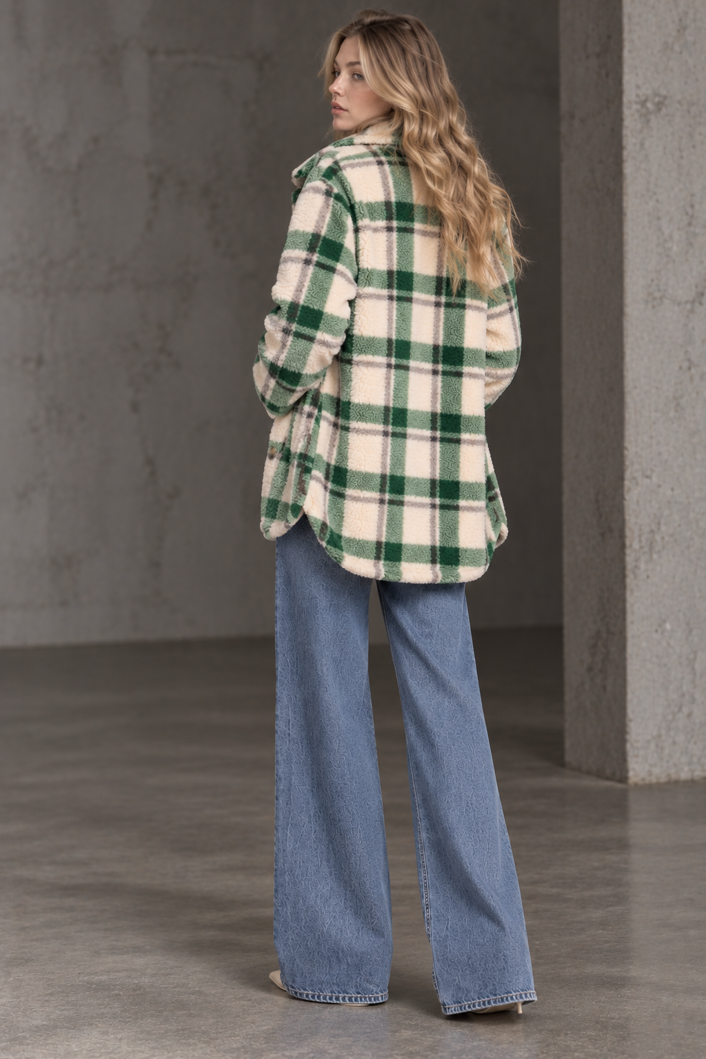A woman stands with her back to the camera wearing an oversized green and cream plaid flannel shirt and wide-leg blue jeans in a concrete-walled room.