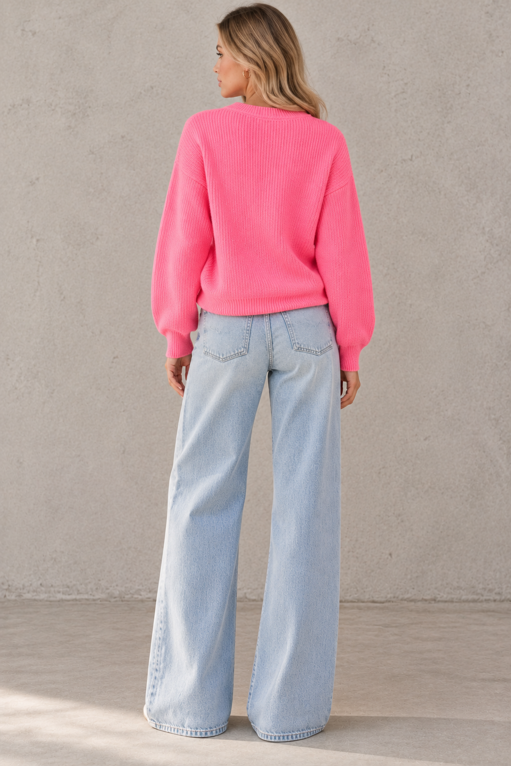 A woman viewed from behind wearing a bright pink ribbed knit sweater and light blue wide-leg jeans, standing against a textured light gray wall.