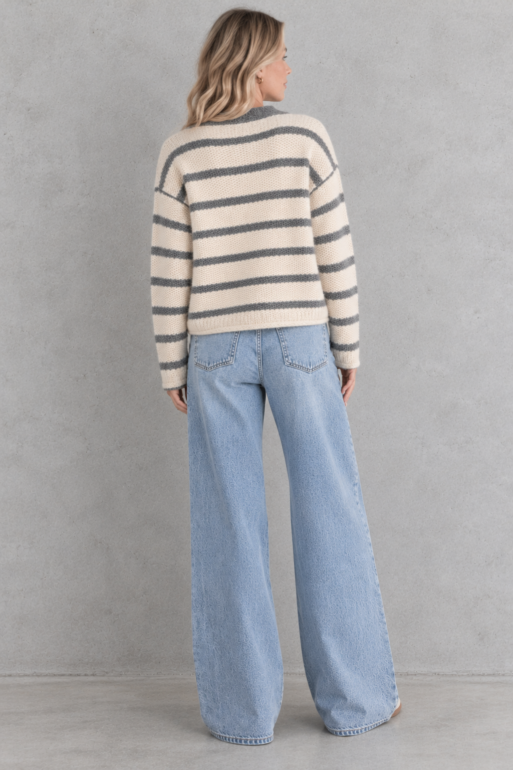 A woman viewed from behind wearing a cream and gray striped knit sweater and light blue wide-leg jeans, standing against a textured gray wall.