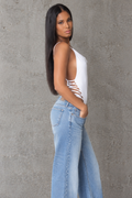 A woman with long dark hair stands in profile against a textured gray concrete wall, wearing a white sleeveless bodysuit with side cutouts and light-wash blue jeans.