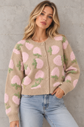 A woman wearing a beige knit cardigan with pink fuzzy floral motifs and green leaf accents, paired with light blue jeans, standing against a textured gray wall.