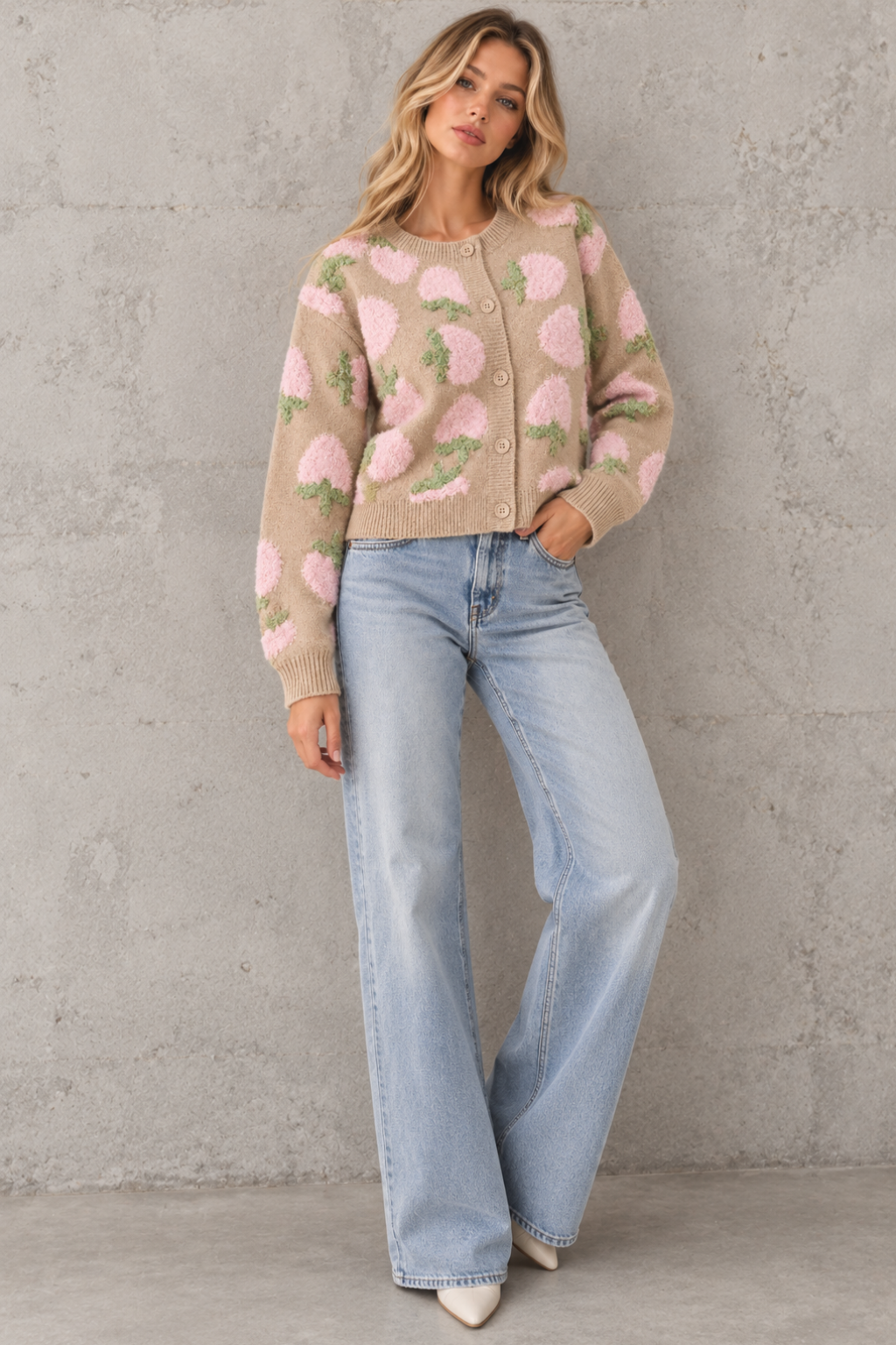 A woman models a beige knit cardigan with a pink and green floral pattern, paired with light-wash wide-leg jeans and white pointed-toe heels, standing against a textured gray concrete wall.