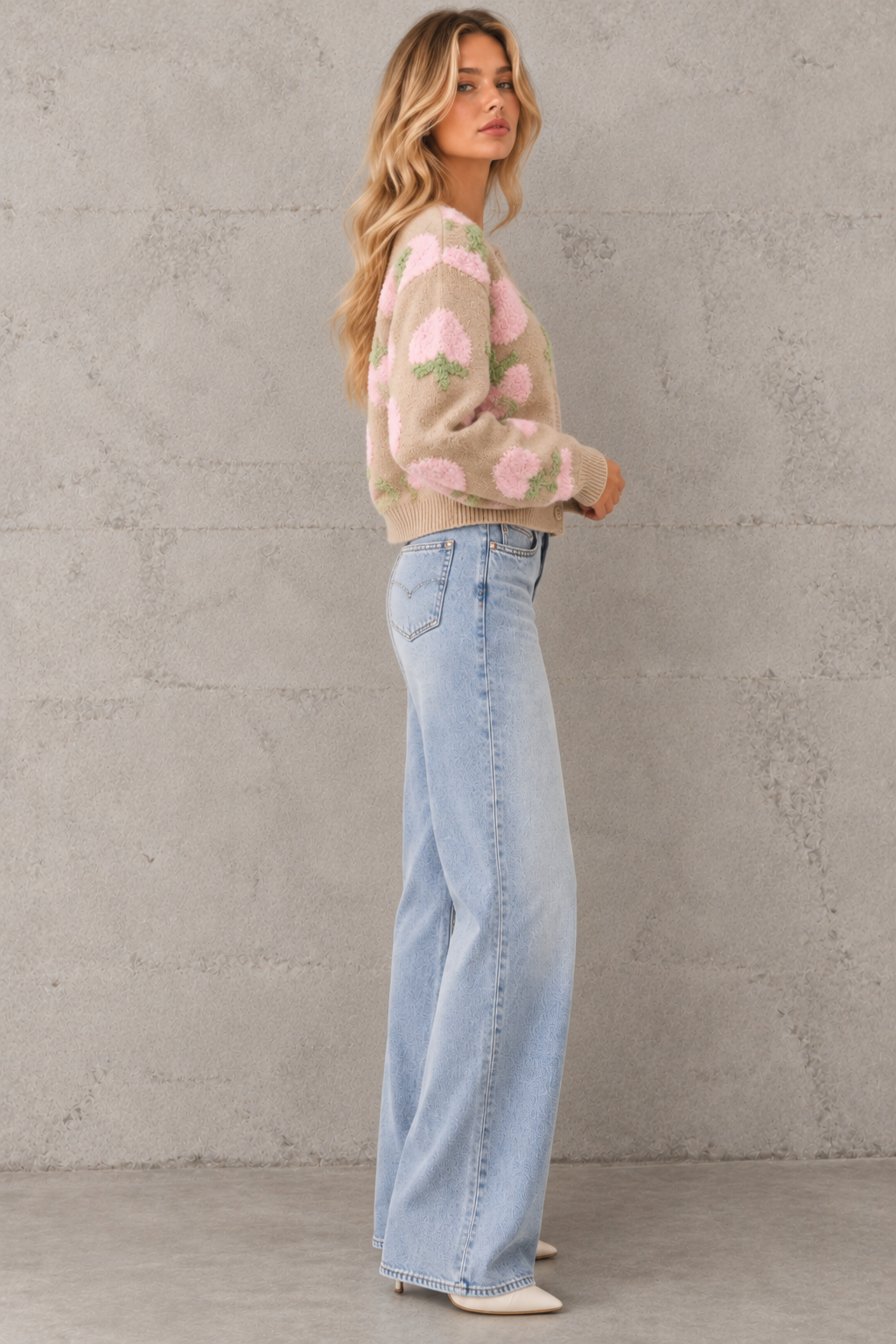 A woman stands in profile wearing a beige knit sweater with large pink tulip-like floral motifs and light blue wide-leg jeans, against a textured gray concrete wall.